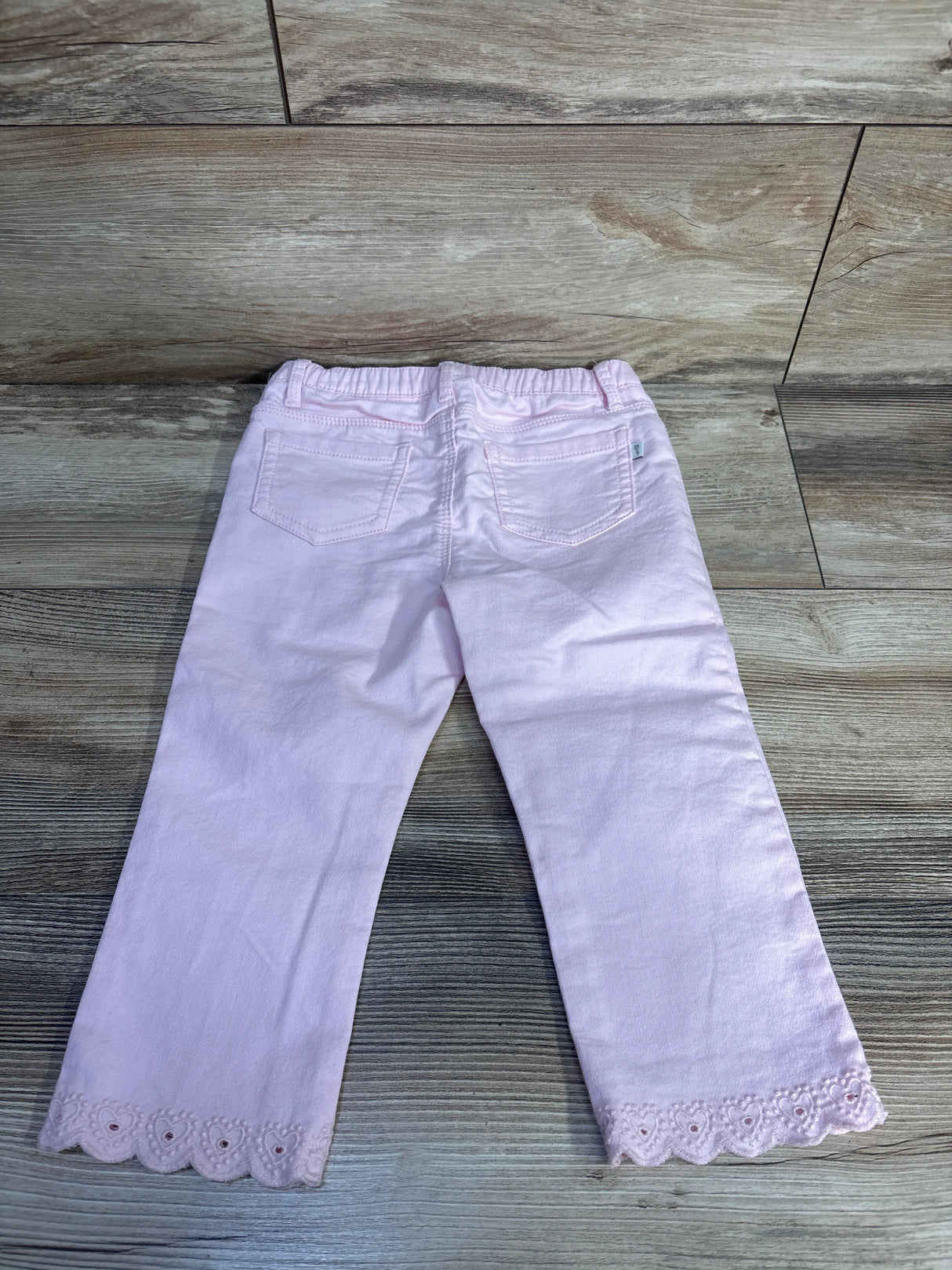 Back view of pink OshKosh toddler jeans (24M) showing scalloped embroidered hem and elastic waistband on wood background