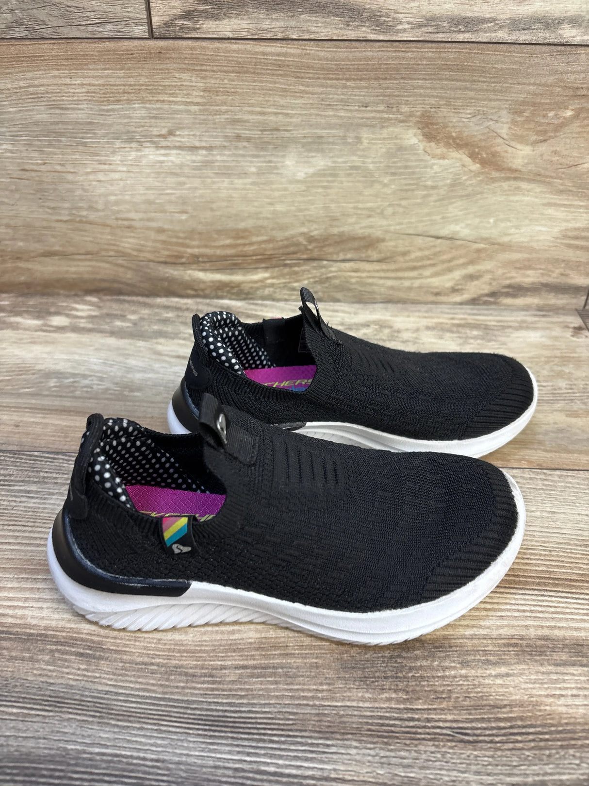 Black slip-on kids sneakers with white soles, polka dot lining, on wooden background