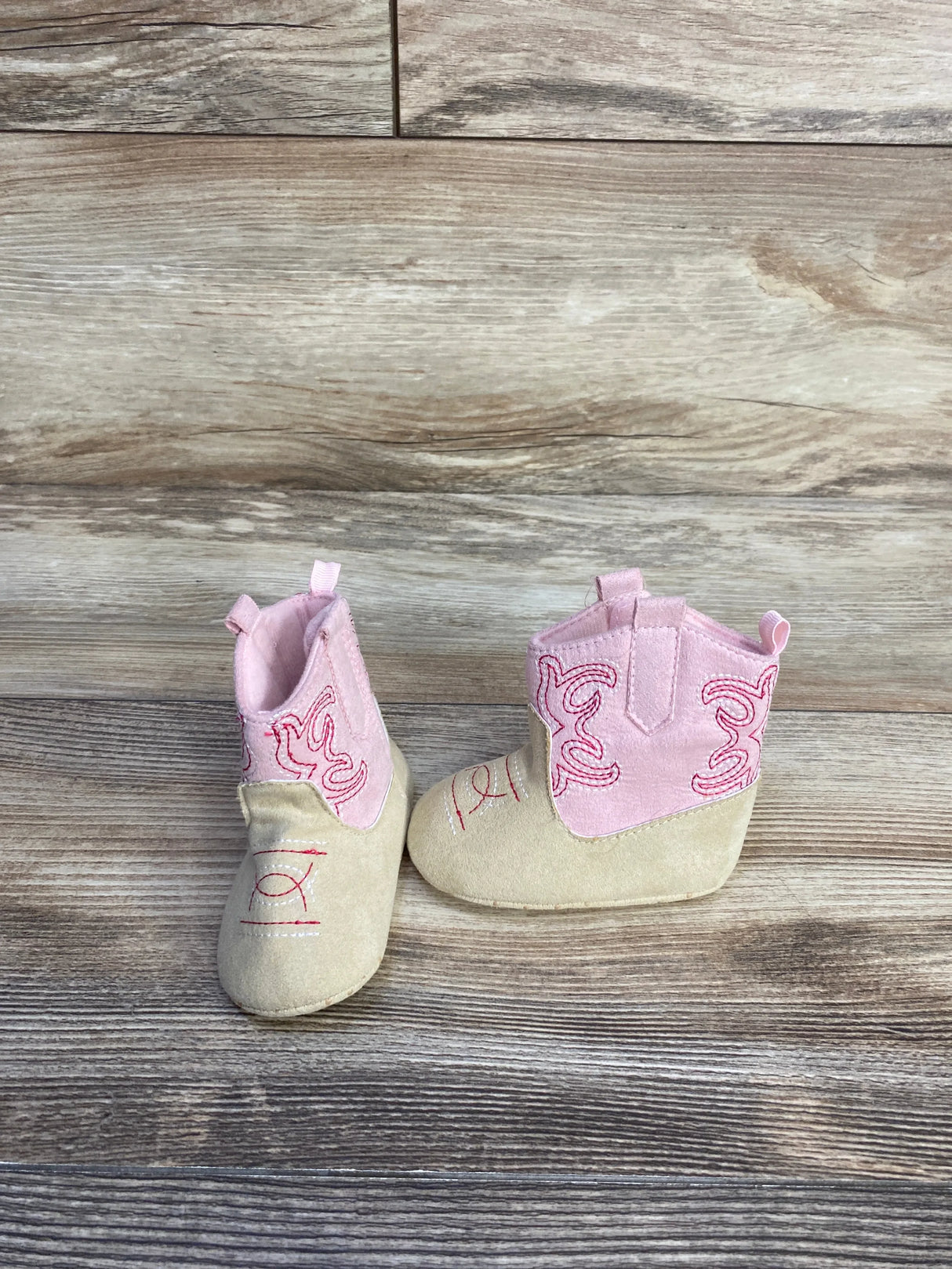 Front and side view of Stepping Stones cowgirl crib boots in pink and tan showcasing embroidered stitching and pull tabs on wood-look background