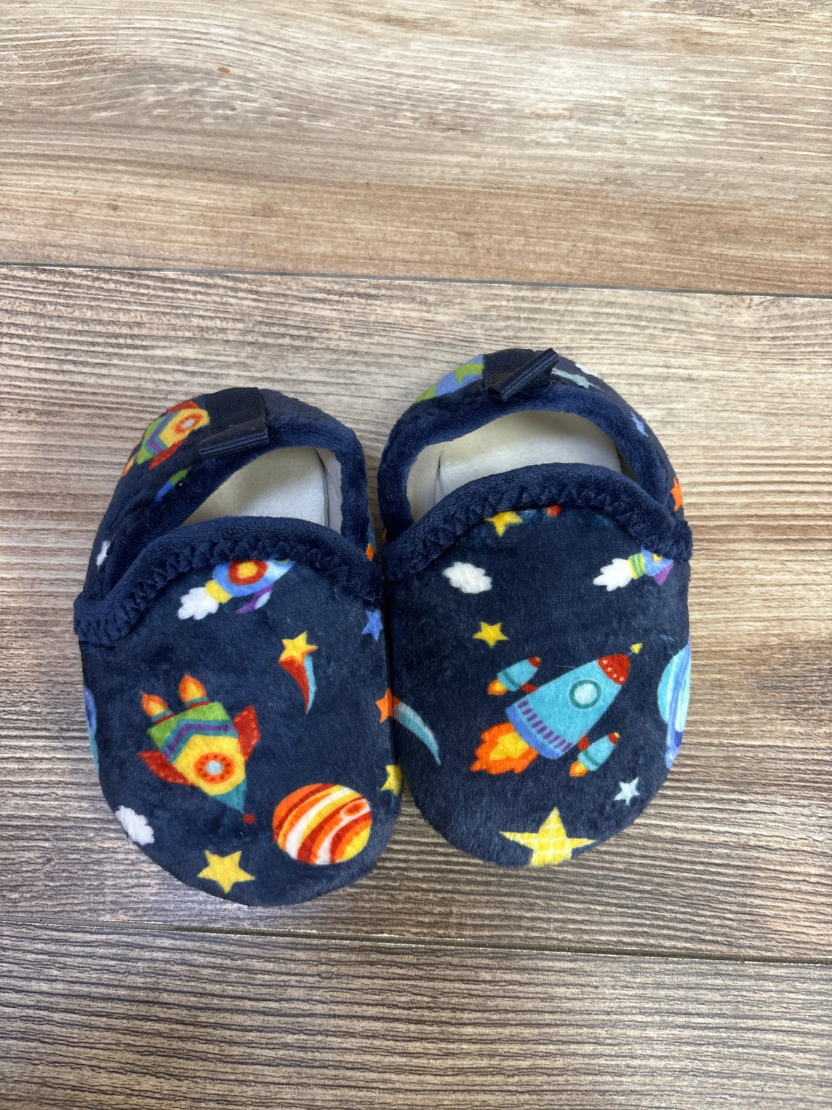 Top-down view of navy baby slippers size 5C showing plush fabric with colorful rocket and planet print and Velcro strap on wood floor
