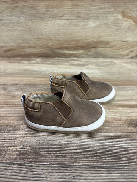 Angled view of brown Robeez Lenny Loafer crib shoes showcasing elastic side panels and white soles on a wood-grain surface