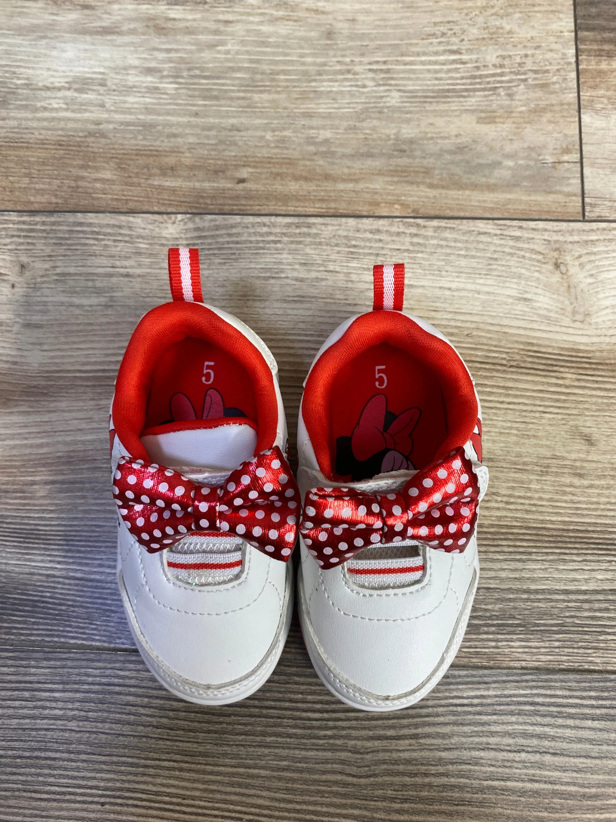 Top-down view of white Minnie Mouse baby sneakers with red polka-dot bows, red lining and visible size 5 on wood floor
