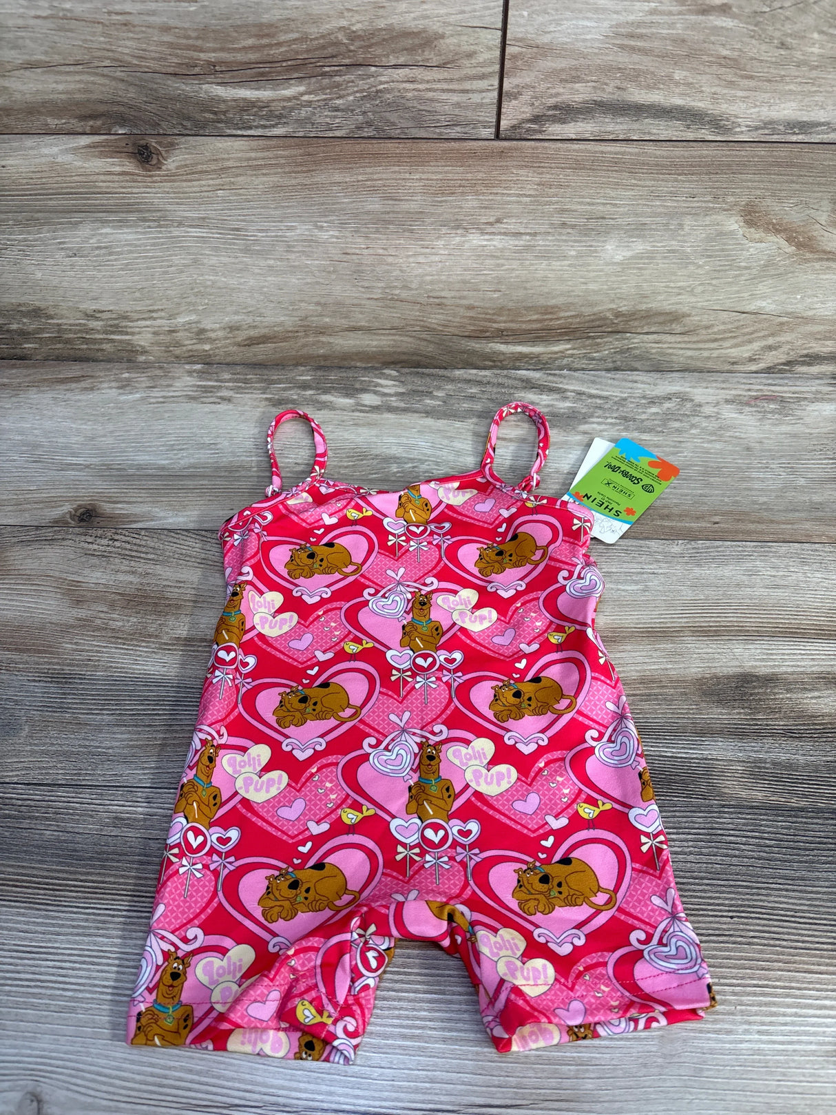 Top-down view of pink Shein x Scooby-Doo romper showing heart and Scooby print, thin straps, and tag on wood background