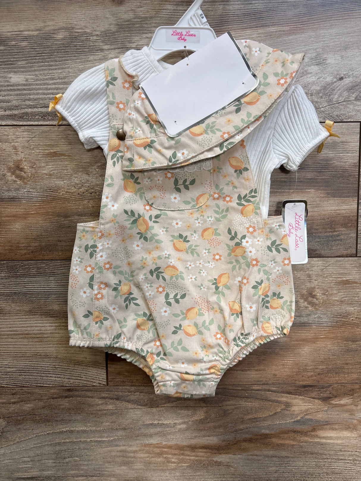 Front view of Little Lass 2-piece: white ribbed shirt with peach floral shortalls and hat on hanger over wood