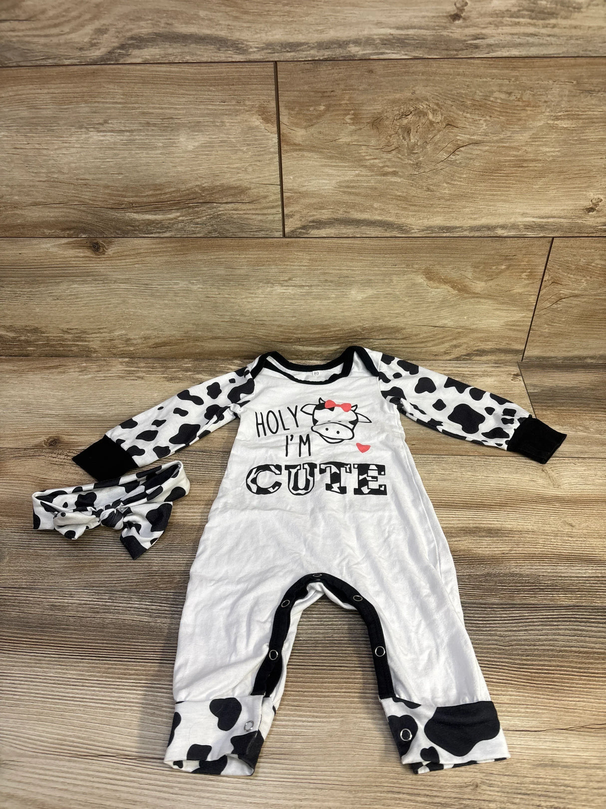 Flat lay white coverall with cow-print sleeves and matching headband, cow graphic reading HOLY IM CUTE, snaps