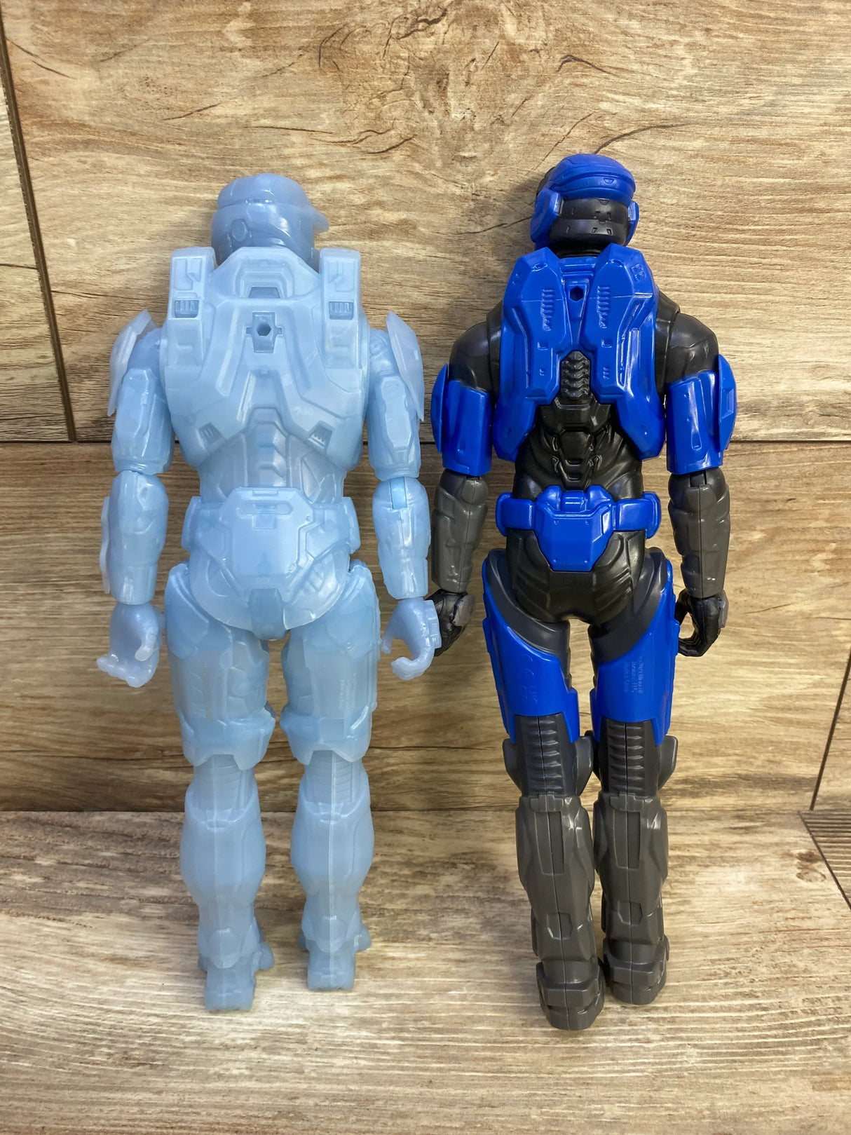 Rear view of two 12-inch Halo Spartan action figures: translucent active-camo and gray figure with blue armor on wood