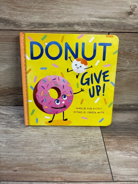 Front of Donut Give Up board book showing yellow cover with pink sprinkled donut character and orange spine on wood floor