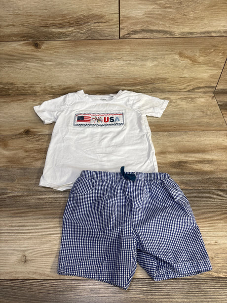 Flat lay of toddler white smocked tee and blue gingham shorts with embroidered USA flag patch on wood floor