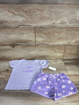 Flat lay of lilac Barbie 2-piece tee and shorts showing logo on tee, lace trim, and floral shorts with bow on wood background