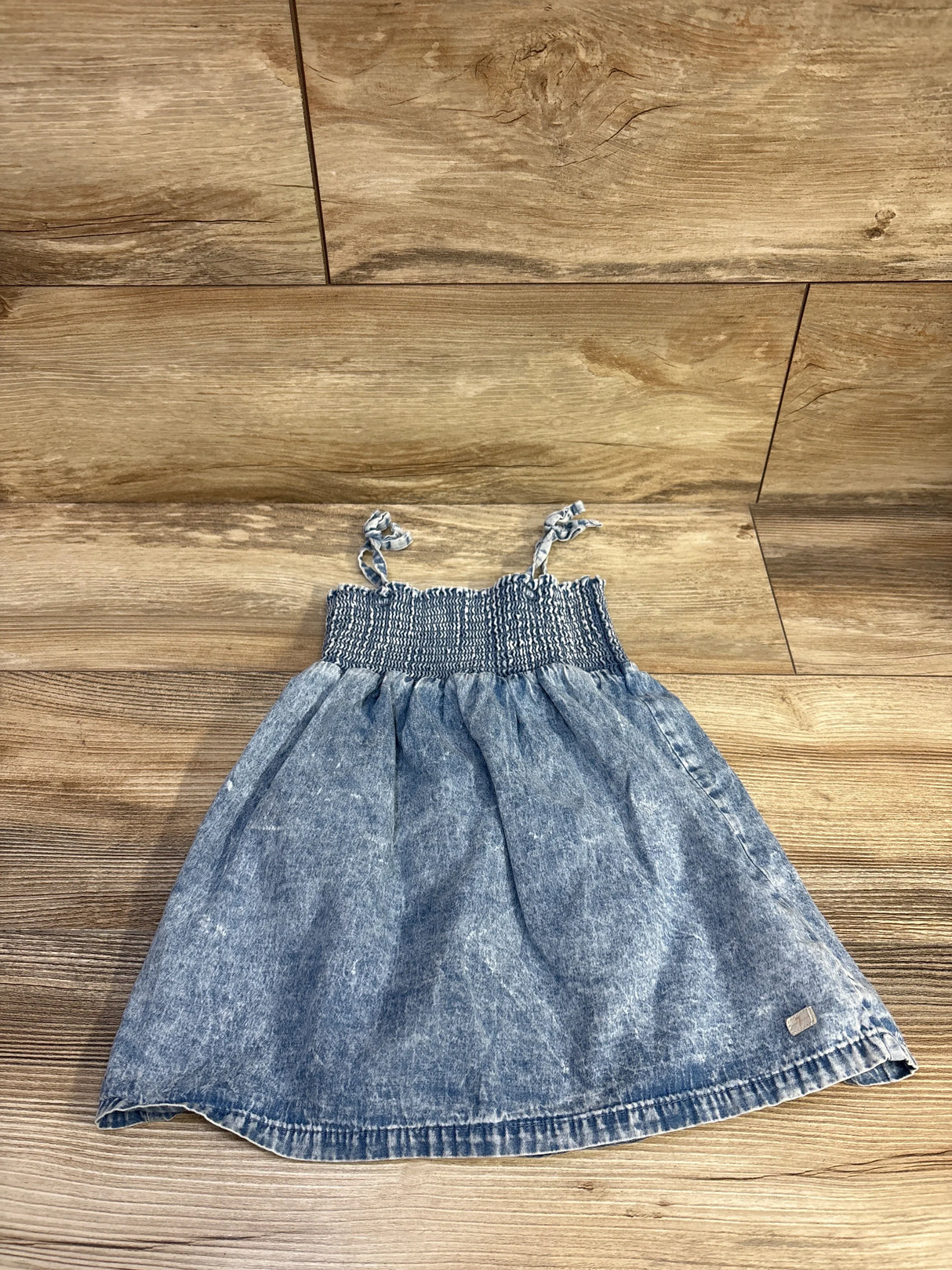 Top down view of blue toddler chambray dress with smocked bodice and knotted shoulder ties on wood floor background