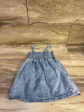 Top down view of blue toddler chambray dress with smocked bodice and knotted shoulder ties on wood floor background