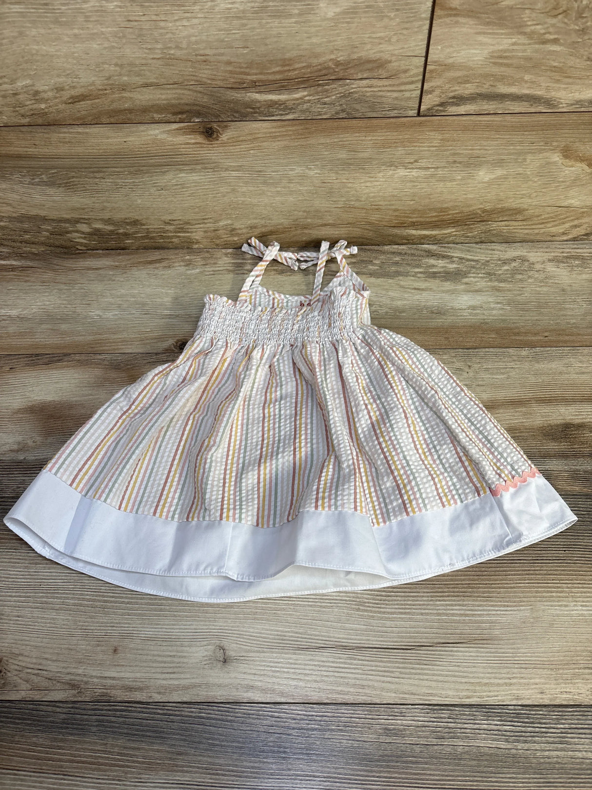 Flat-lay of Goodlad 2T embroidered giraffe gingham dress with smocked bodice, tie straps, pastel stripes on wood background