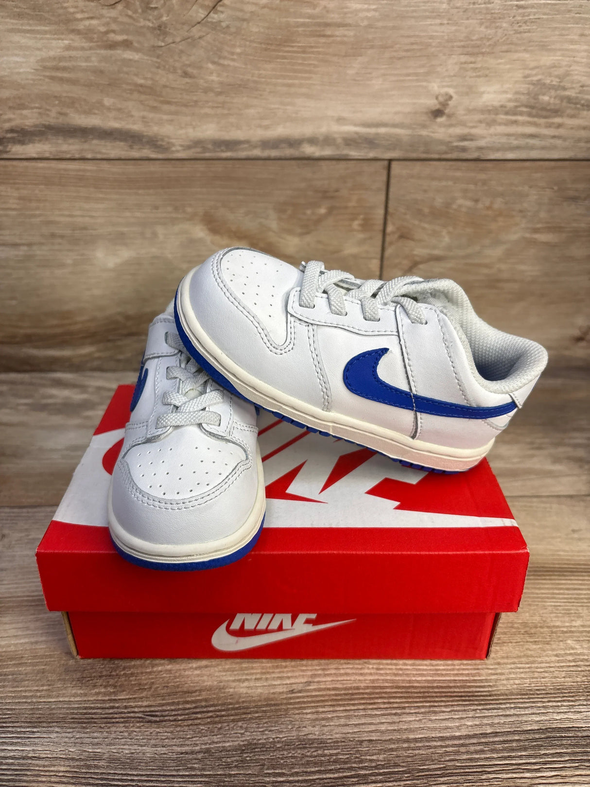 Three-quarter view of Nike Dunk Low TD toddler sneakers size 8C in Summit White with Hyper Royal Swoosh and blue sole on red Nike box, wood background