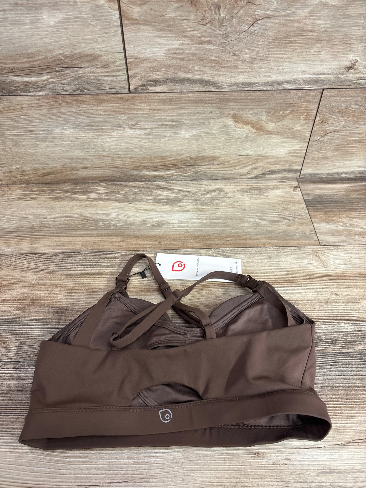 Flat lay of espresso nursing and pumping sports bra showing wide underband with pump access, crossed adjustable straps, on wood floor