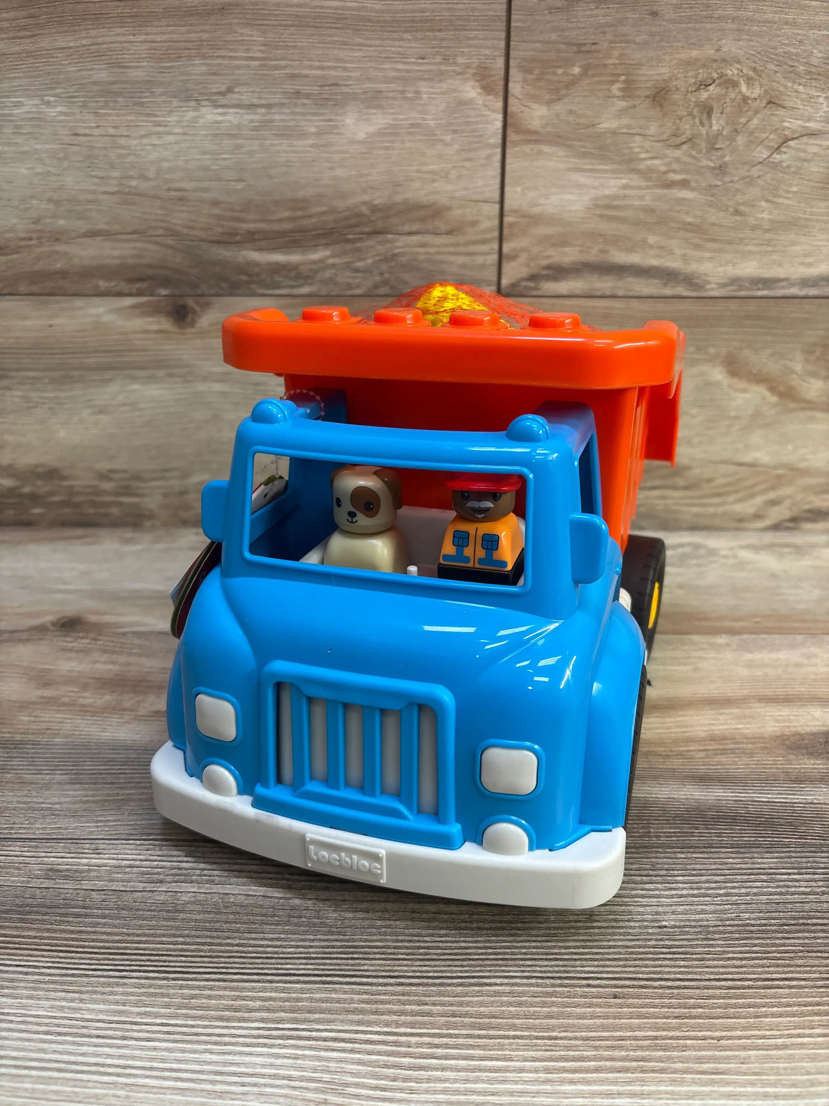 Front view of Battat dump truck with building blocks, blue cab, orange dump bed, and two figures on a wood-grain background