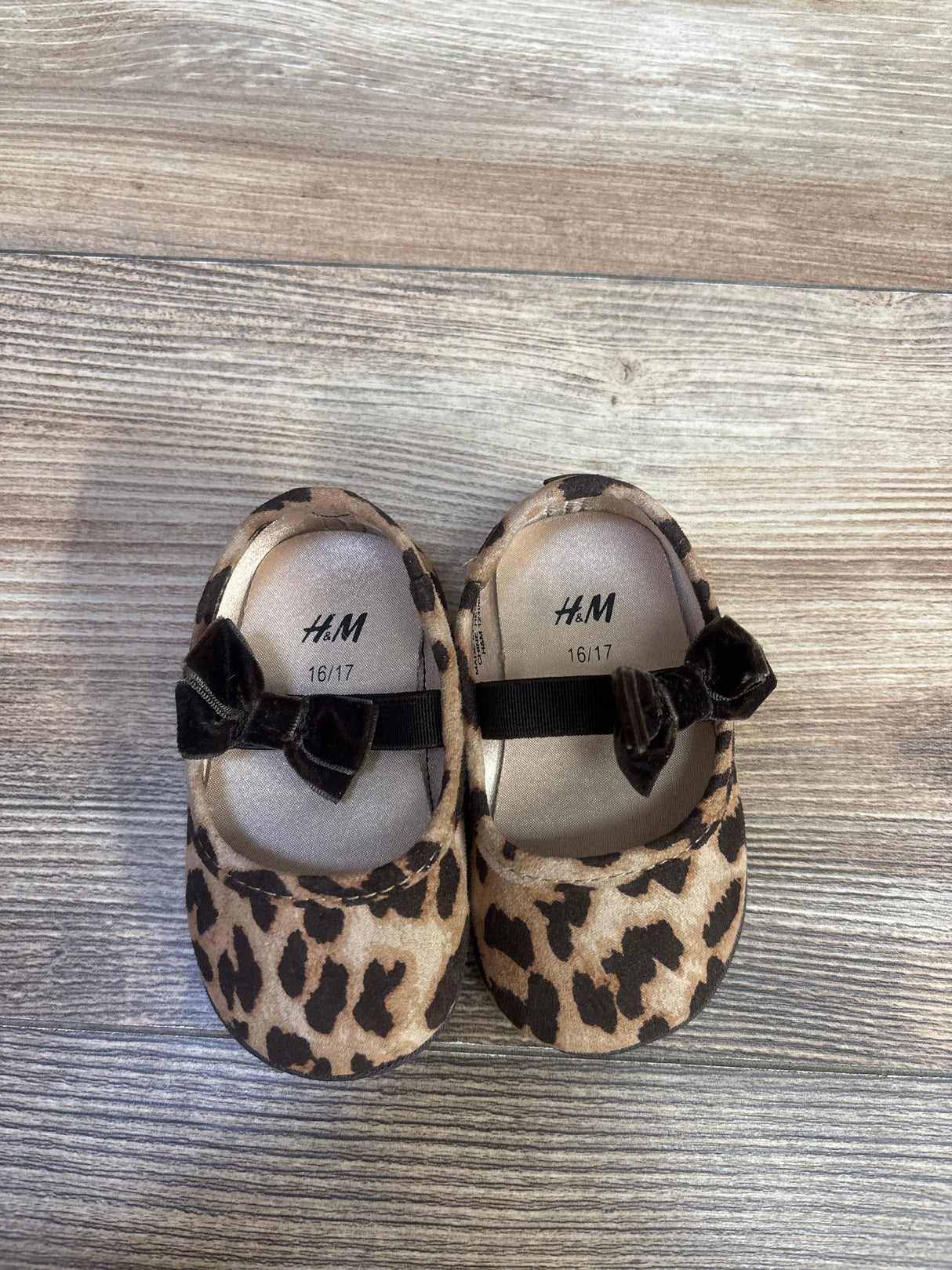 Top-down view of H&M infant leopard-print ballet flats with velvet bows on light wood floor, insoles marked 16/17