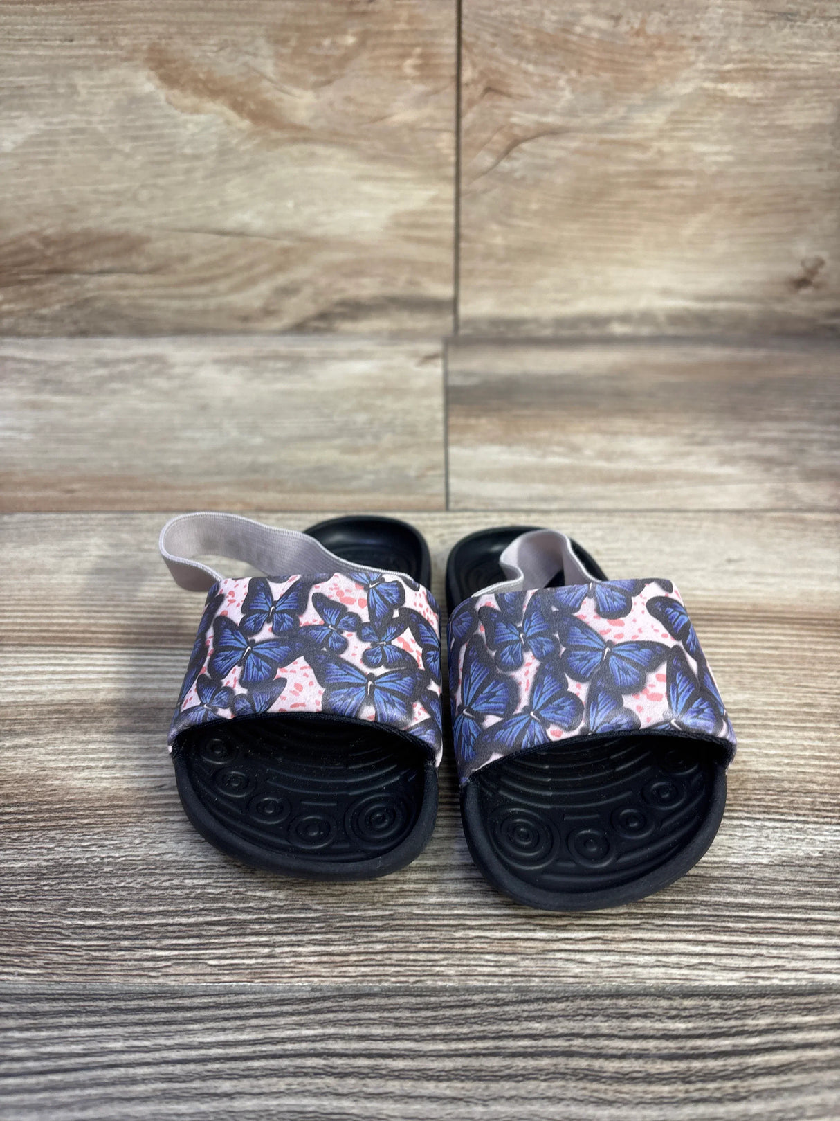 Front view of Nike Kawa Slide SE toddler sandals showcasing pink butterfly strap and black contoured soles on wood-look floor
