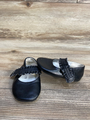Pair of black baby shoes with ruffled straps on wood grain surface