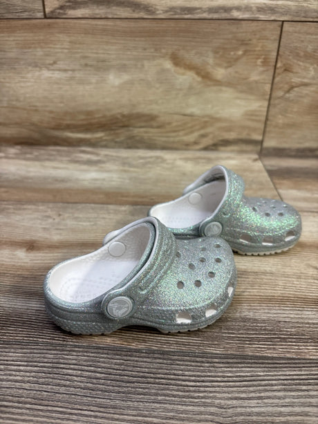 Angled view of Crocs Classic Glitter Clogs in silver showcasing glitter finish, ventilation holes, and heel straps on wood flooring