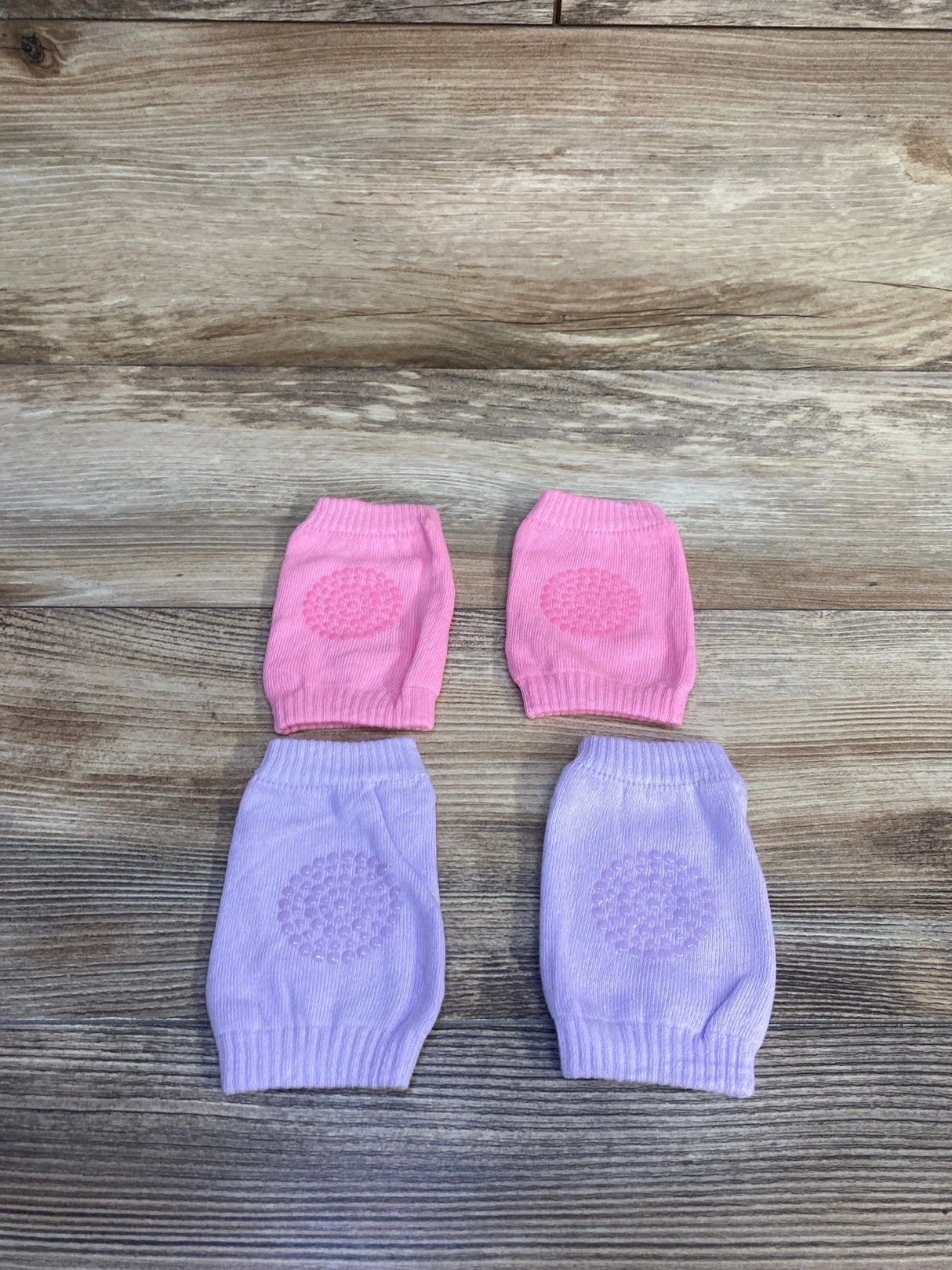 Top view of two pairs of baby crawling knee pads with textured non slip grip on pink and lilac knit, on wood background