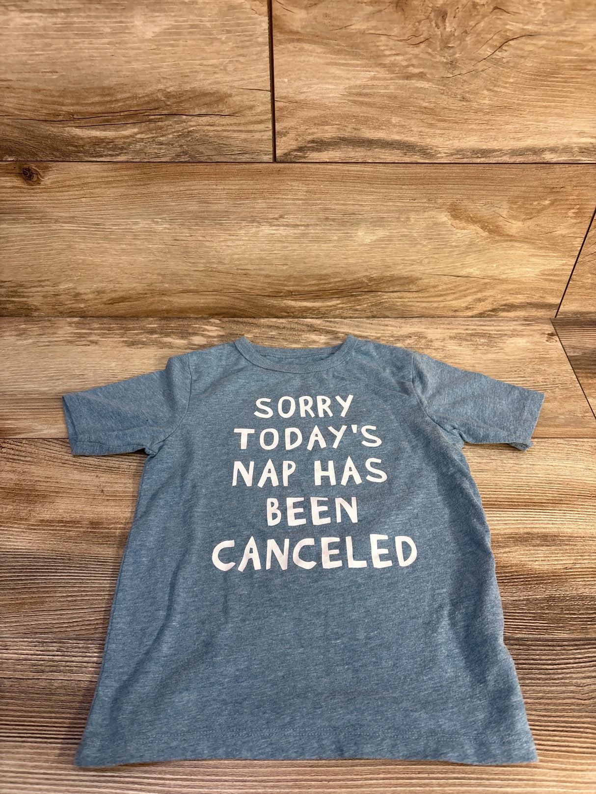 Flat lay of blue Children’s Place 4T T-shirt showing white text "SORRY TODAY'S NAP HAS BEEN CANCELED" on wood floor background
