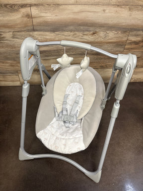Overhead of Graco Slim Spaces baby swing showing padded seat, harness, and two hanging star toys on wood-paneled backdrop