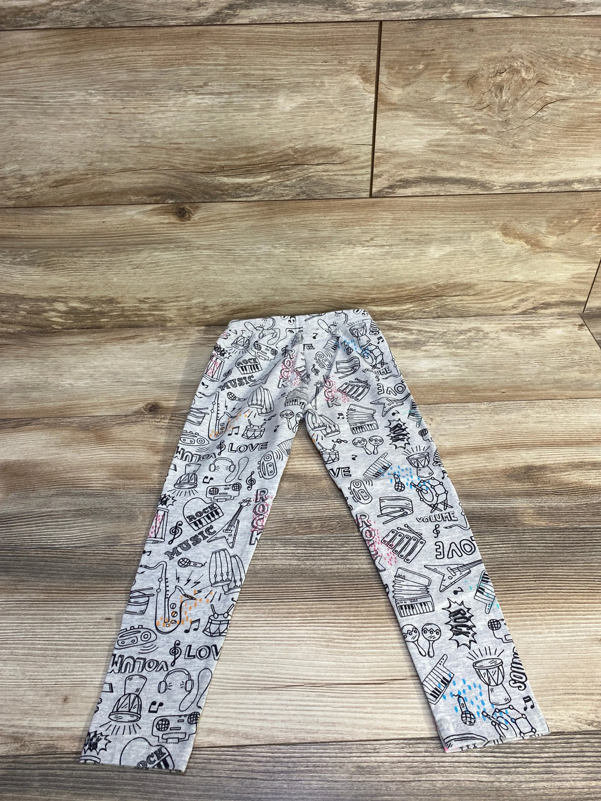Flat lay of grey toddler leggings showcasing black music-themed doodle print with small pink and blue accents on a wood floor background