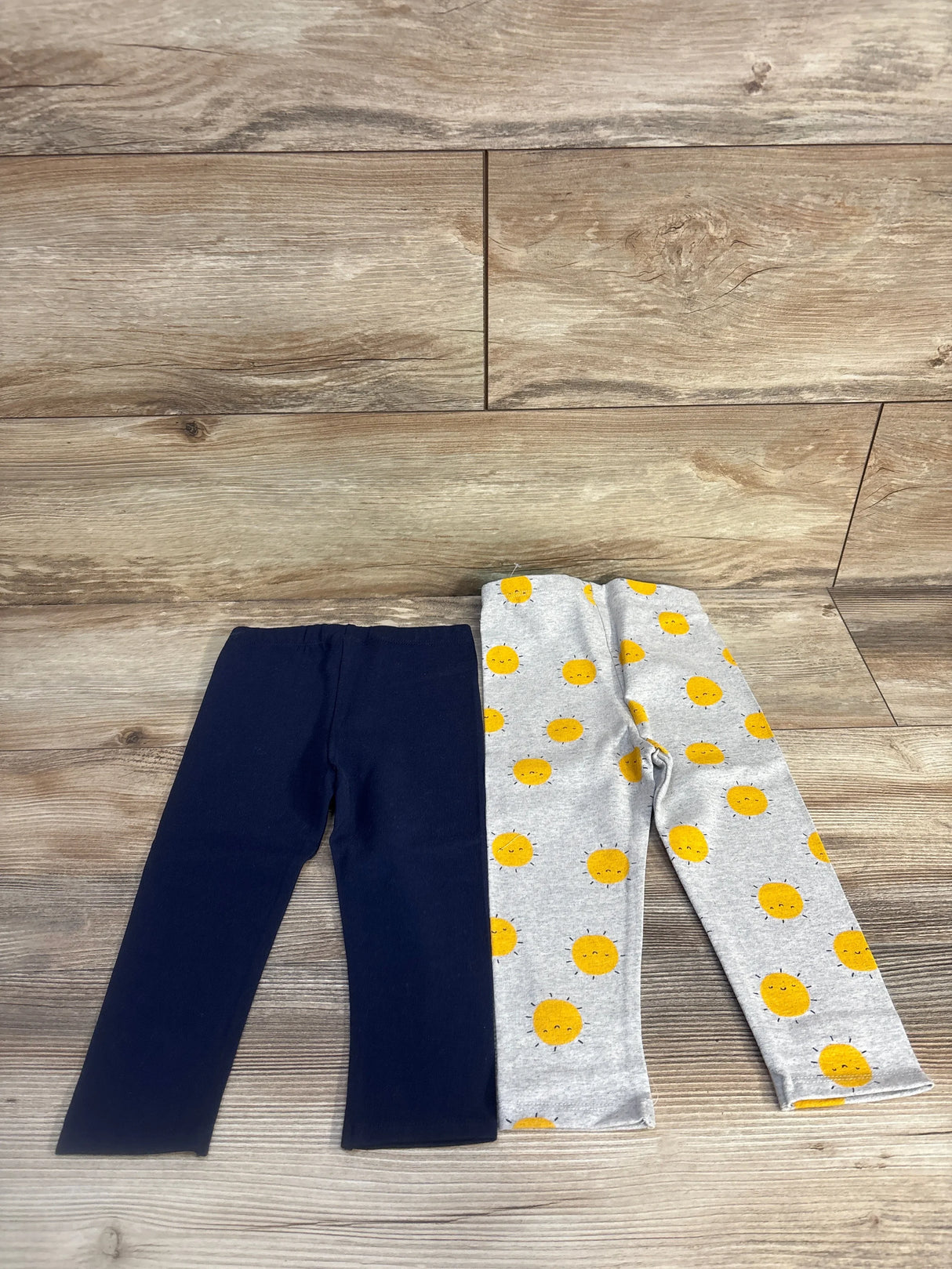 Flat lay of two 18-month girls leggings: one solid navy and one grey with yellow sun faces, elastic waist on wood background