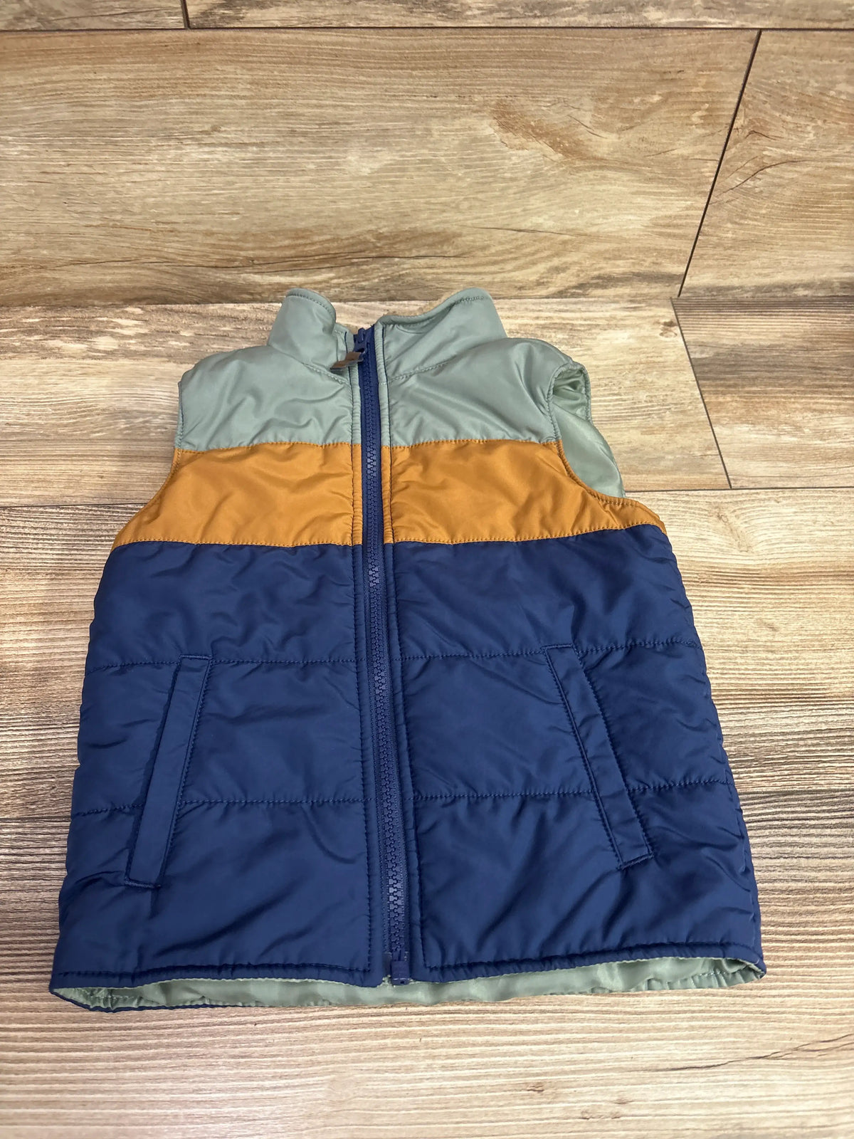 Front view of Carter’s full-zip vest jacket in green, mustard, and navy showcasing quilted panels and zippered pockets on a wood floor