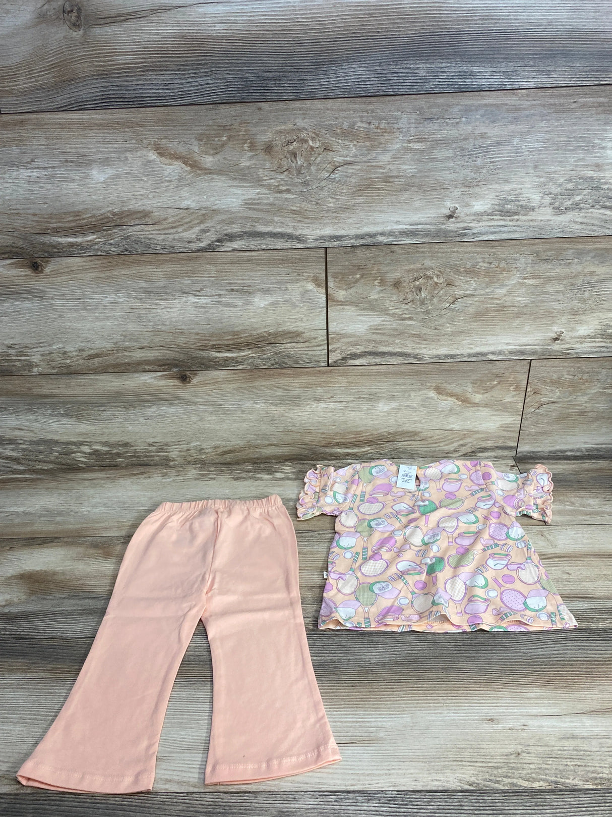 Peach-colored toddler pants with elastic waist and pastel patterned short-sleeve top on wood floor