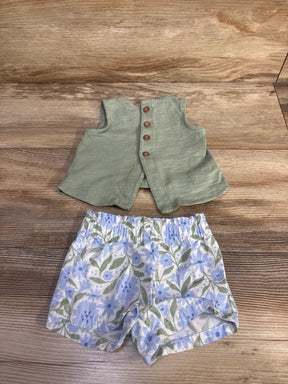 Top-down view of 2-piece baby outfit: sage sleeveless button-back top and white shorts with blue floral print on wooden background