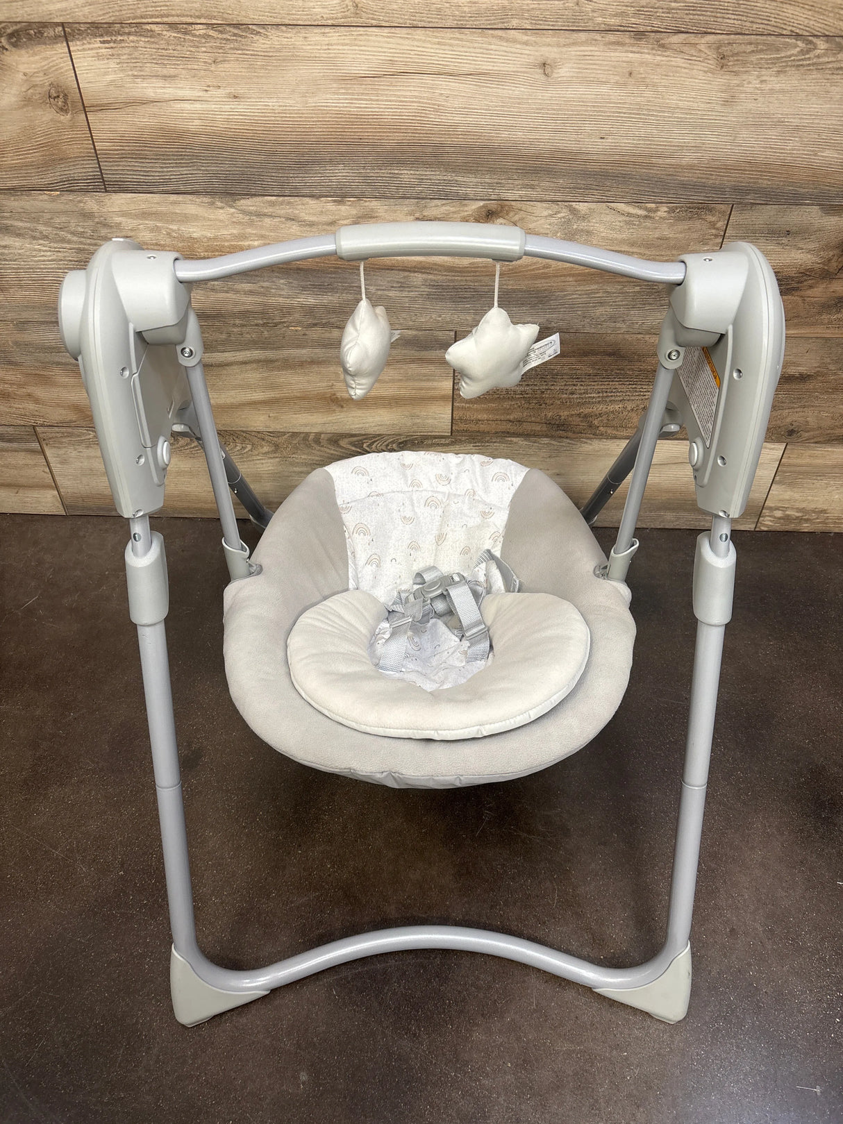 Front view of Graco Slim Spaces compact baby swing showing padded light-gray seat with infant head support, hanging plush toys, and foldable silver frame against a wood-paneled backdrop