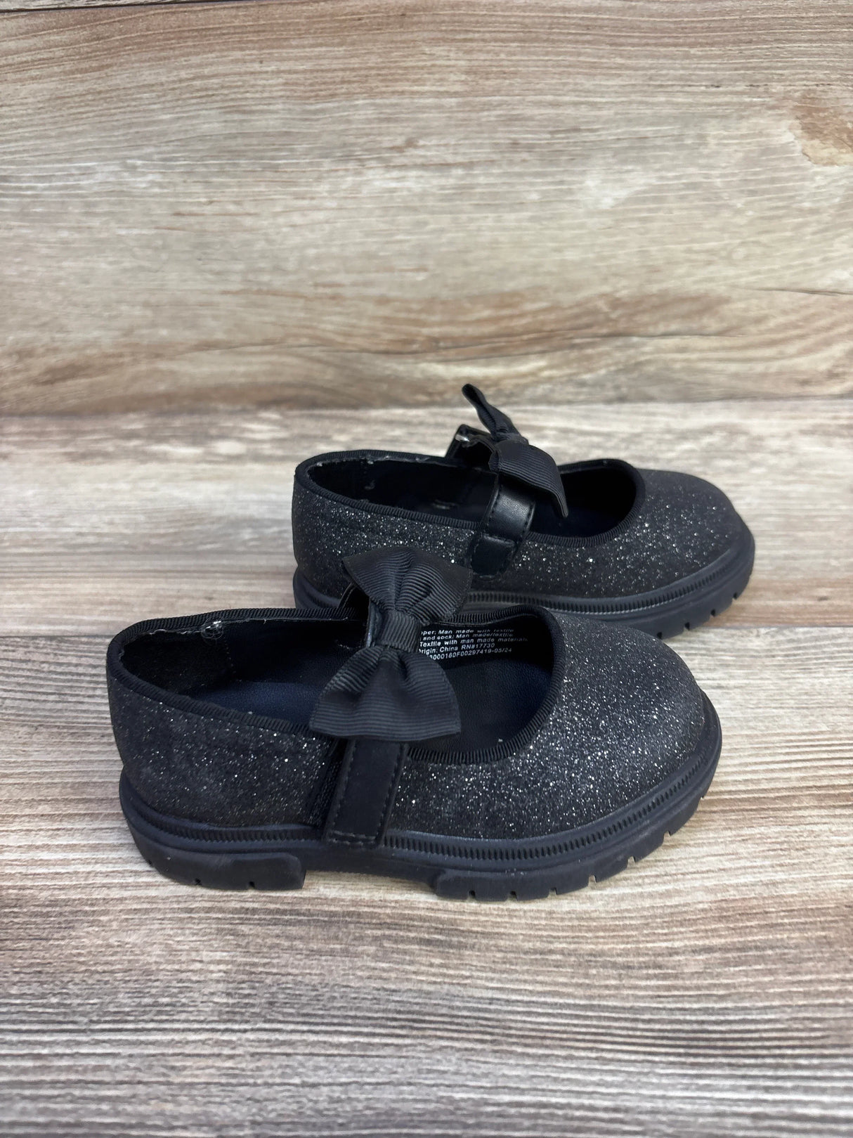 Side view of Cat & Jack Kori Mary Jane flats showcasing black glitter upper, bow strap, and lug sole on wood plank background