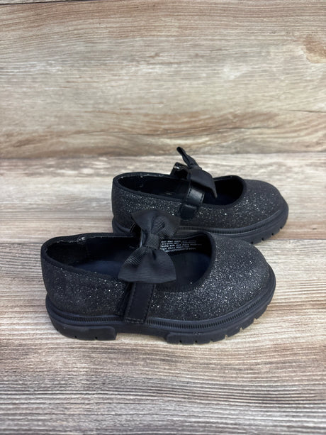 Side view of Cat & Jack Kori Mary Jane flats showcasing black glitter upper, bow strap, and lug sole on wood plank background