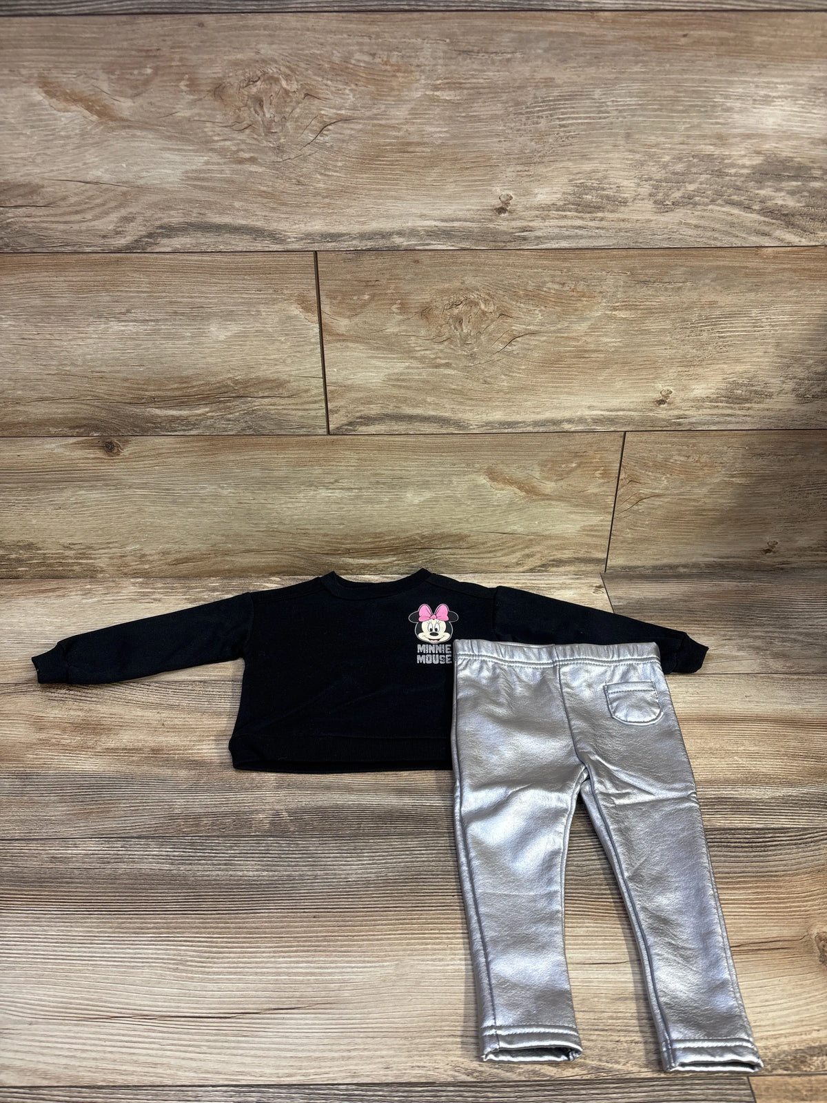 Top-down view of Disney Baby Minnie Mouse sweatshirt and leggings set showcasing a black sweatshirt and silver leggings on a wood floor