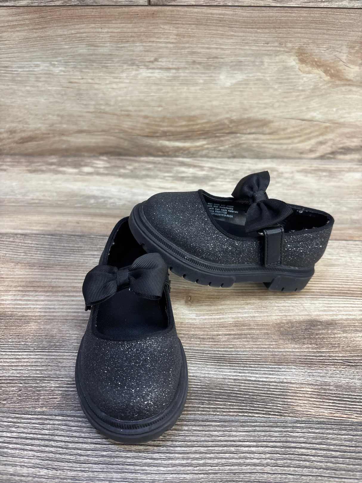 Front and side view of Cat & Jack Kori Mary Jane flats, black glitter, showing lug sole tread and grosgrain bow on light wood