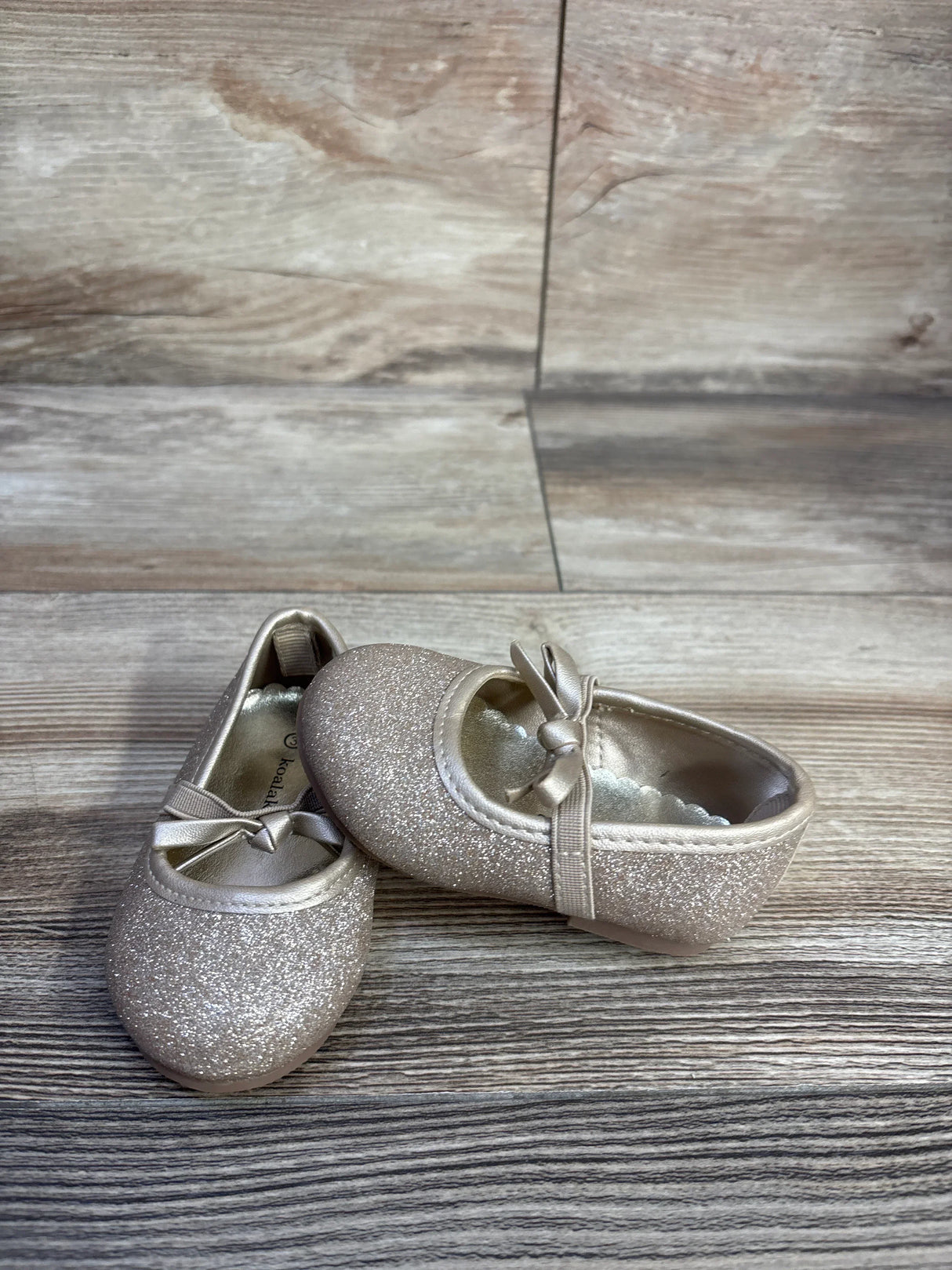 Angled front view of Koala Kids gold glitter bow ballet flats, size 3C, showing glitter finish and ribbon bow on wood background