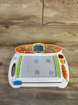 Front view of VTech Write & Learn showing magnetic drawing board, orange tethered stylus, and control panel screen