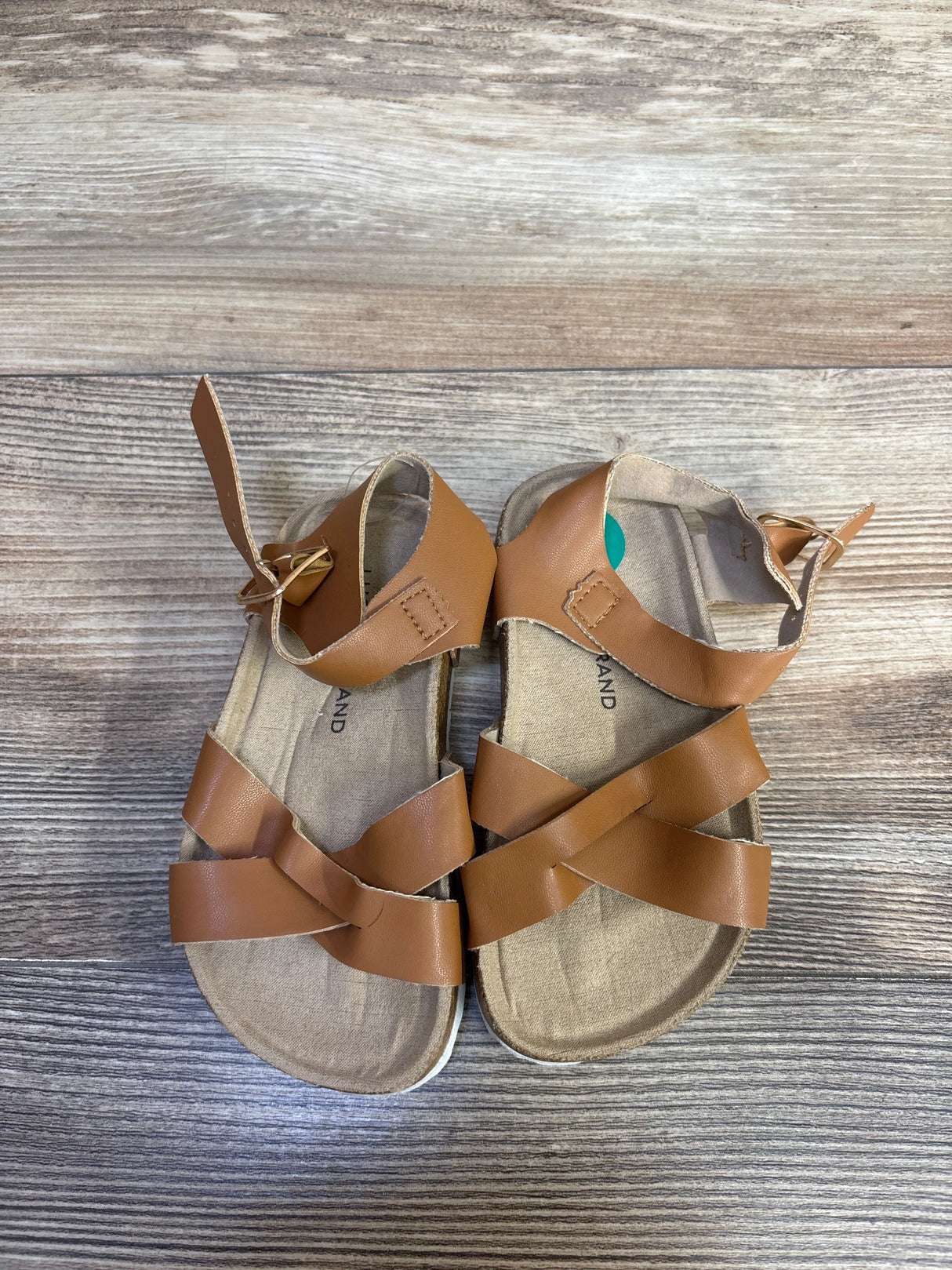 Top down view of Lucky Brand Kiara toddler sandals in brown showcasing crisscross straps and ankle buckle with wood plank background