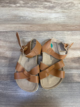 Top down view of Lucky Brand Kiara toddler sandals in brown showcasing crisscross straps and ankle buckle with wood plank background