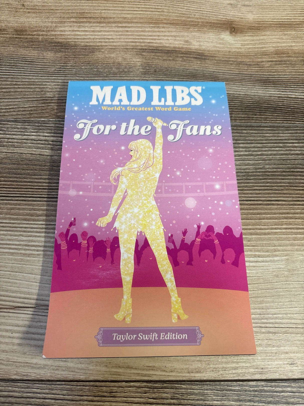 Top-down front view of Mad Libs For the Fans Taylor Swift Edition cover showing gold singer silhouette with pink concert crowd on wooden table