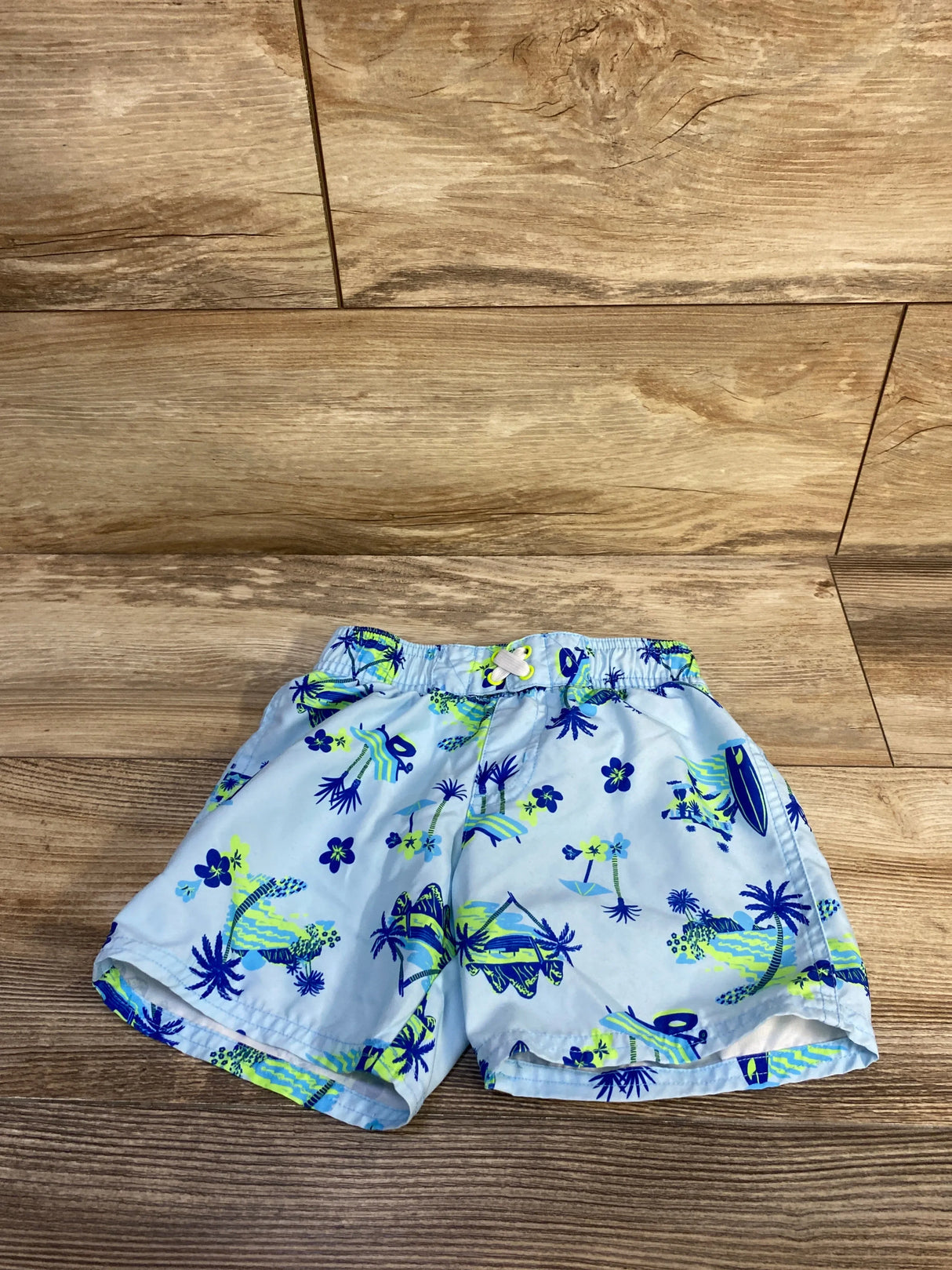 Front view of blue 4T Cat and Jack swim trunks with neon green tropical print and elastic drawstring on wood background