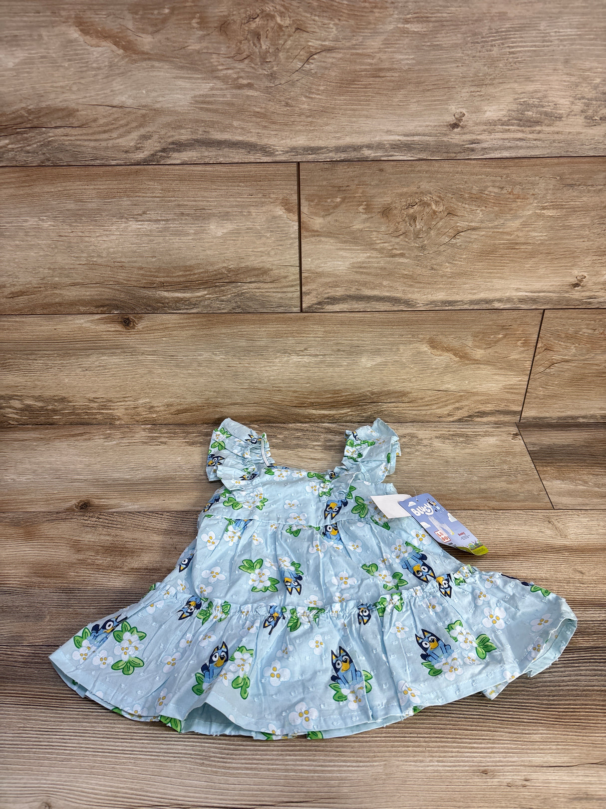 Flat-lay front of light-blue Bluey swiss-dot baby dress showing ruffled sleeves and Bluey floral print on wood background