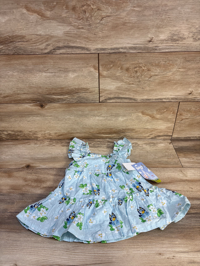 Flat-lay front of light-blue Bluey swiss-dot baby dress showing ruffled sleeves and Bluey floral print on wood background
