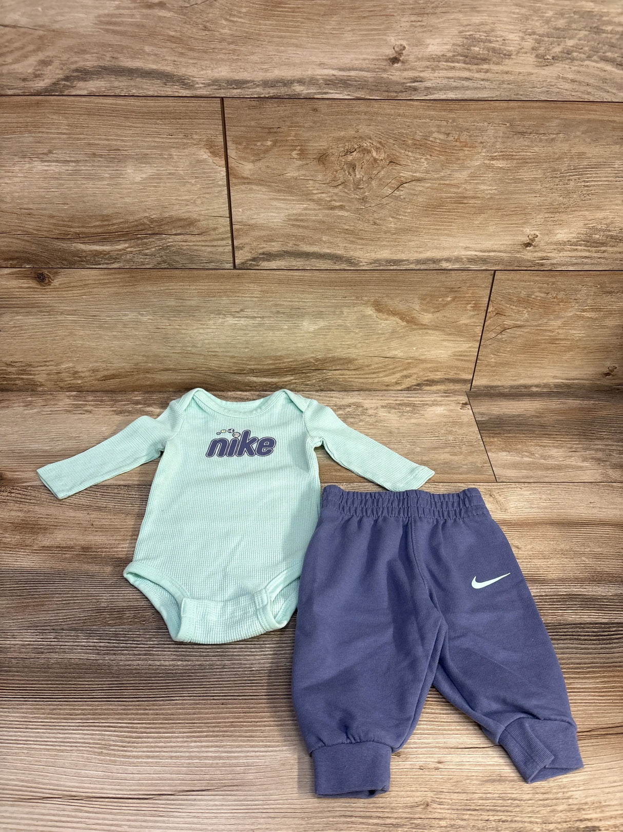 Flat lay of Nike 2-piece bodysuit and joggers showing Nike chest logo on light blue top and purple joggers on wood background