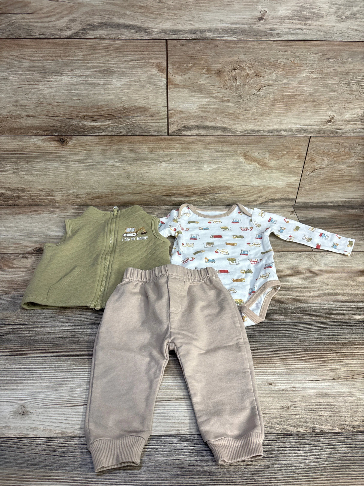 Top view of 3-piece baby set I Dig My Mommy: green quilted vest, vehicle-print long-sleeve bodysuit, beige joggers on wood background