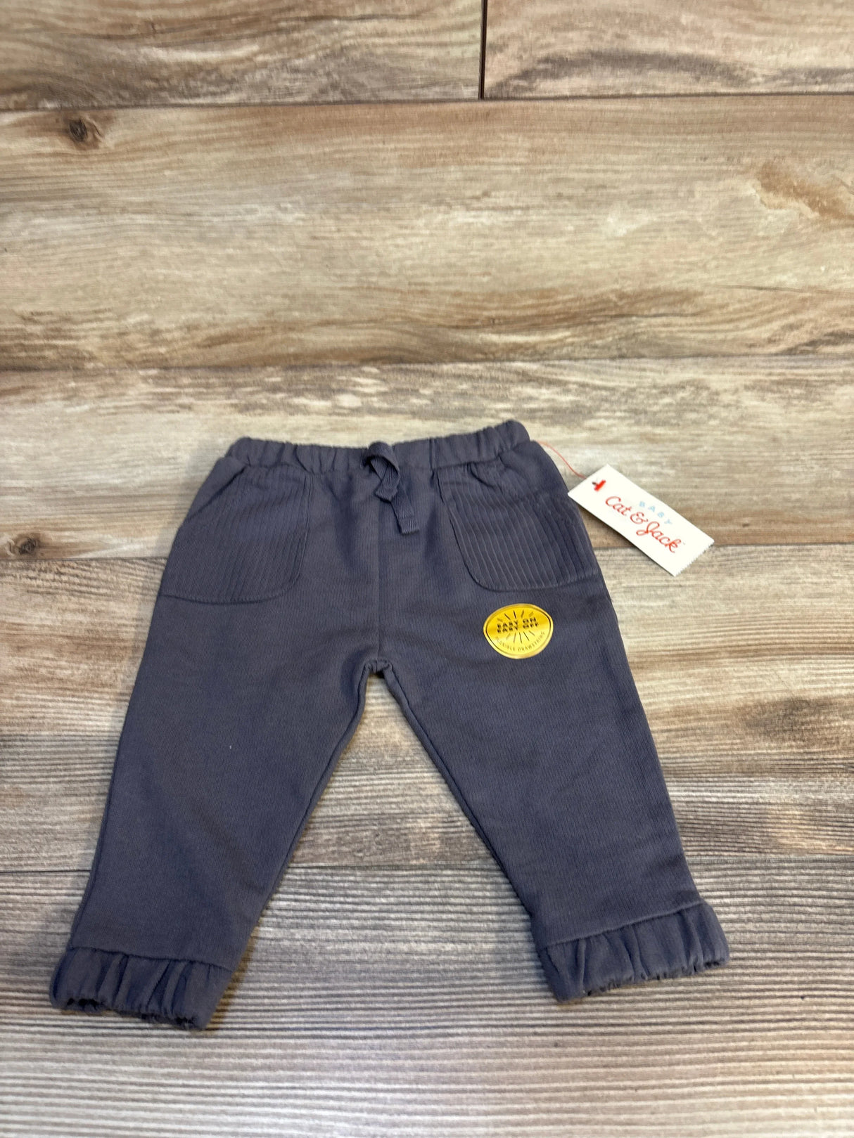 Gray baby pants with ruffled cuffs, Cat & Jack tag, and yellow 'Baby Sun' patch on wood floor