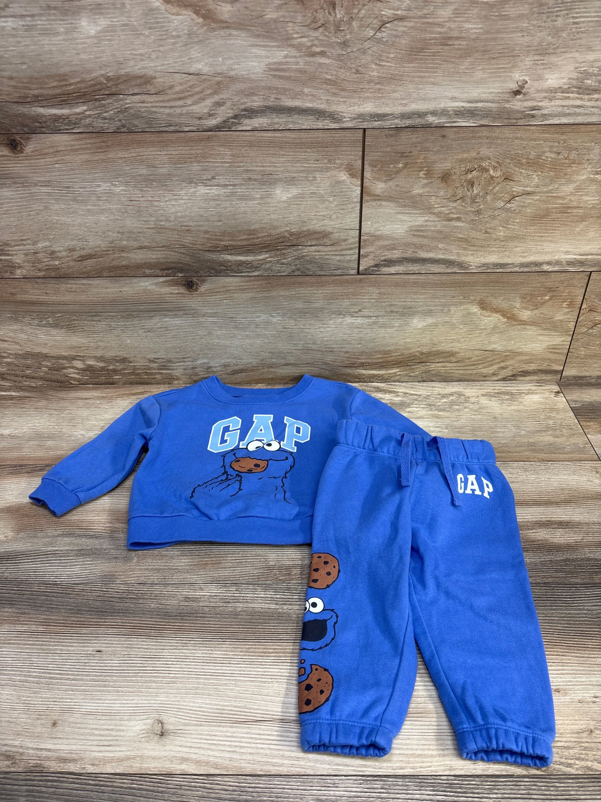 Flat lay of blue Baby Gap Sesame Street 2pc set: Cookie Monster sweatshirt and matching joggers with cookie graphics on wood