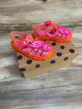 Three-quarter view of Sophia Webster toddler jelly sandals showcasing pink butterfly appliques and orange soles on a polka-dot shoebox background