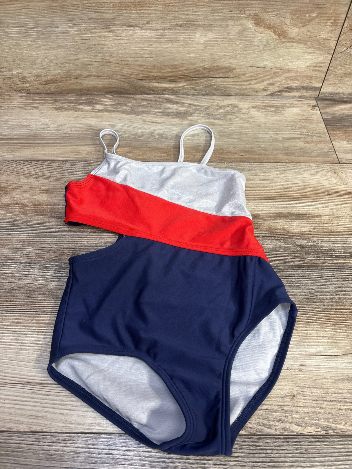 Flat lay of girls red white blue one-piece cut-out swimsuit size 5T showing diagonal red band, silver top, navy bottom, and thin shoulder straps on wood floor background